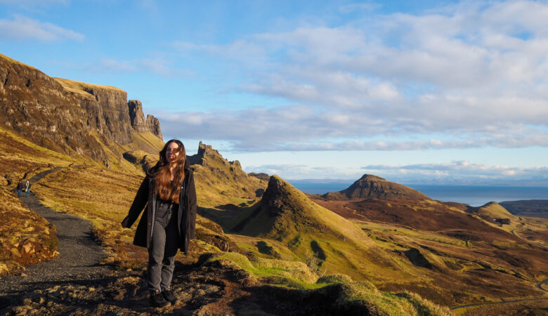 A woman with long hair stands on a rocky path in a hilly, grassy landscape under a blue sky with scattered clouds, with dramatic cliffs and distant hills in the background.