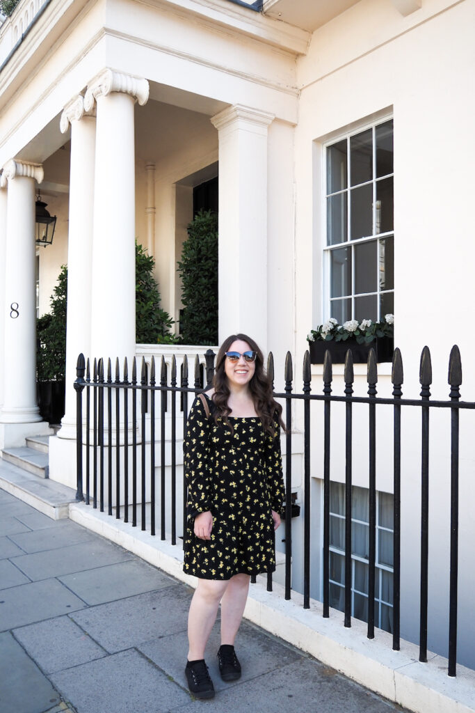 A woman with long brown hair wearing sunglasses and a black dress with yellow floral patterns stands smiling on a sidewalk in front of a white building with columns and black railings.