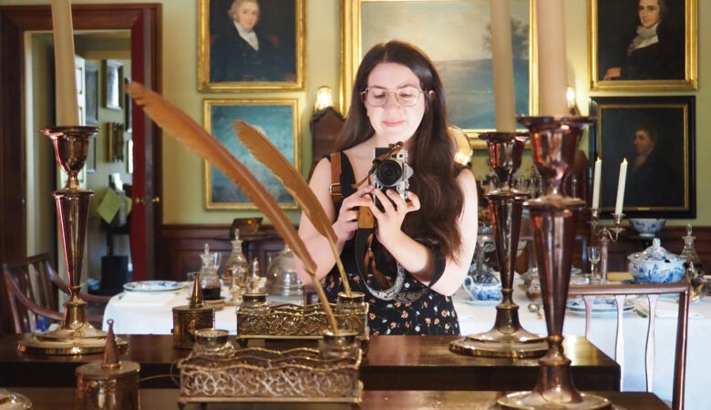 A woman with long hair and glasses takes a photo in a vintage room, reflected in a mirror. Antique candlesticks, quills, and ornate decor surround her, with paintings and a set dining table in the background.