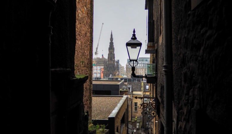 A view of the Scott Monument in Edinburgh, framed by the dark stone walls of a narrow alleyway with a hanging street lamp in the foreground.