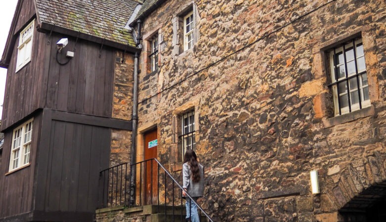 A person walking up stone steps toward the entrance of an old building constructed of dark wood and rough stone.
